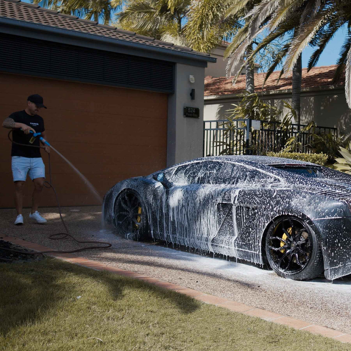 Person washing a black sports car with soap suds in a driveway.