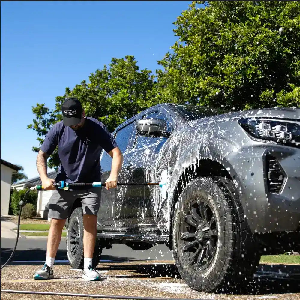 Person washing a gray SUV with a pressure washer on a sunny day.