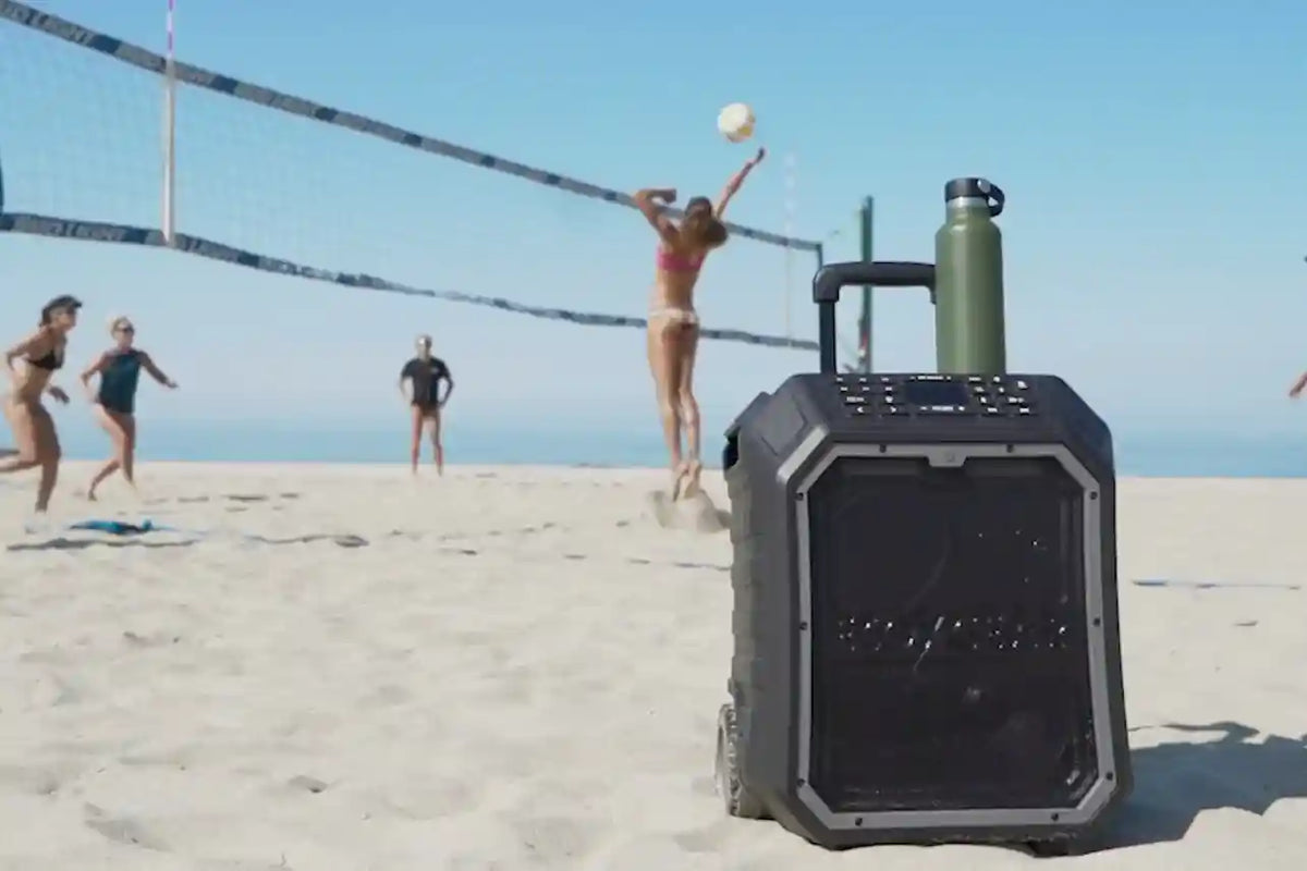 Portable speaker on a sandy beach with people playing volleyball in the background