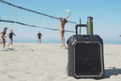 Portable speaker on a sandy beach with people playing volleyball in the background