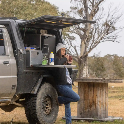 Person sitting in an open truck bed with a portable kitchen setup in a natural setting.