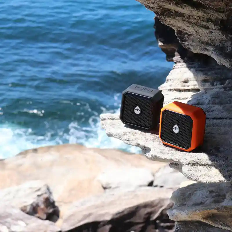Two portable speakers, one black and one orange, on a rocky surface with ocean in the background.