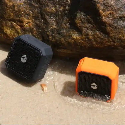 Two portable speakers, one black and one orange, on a sandy surface with rocks in the background.