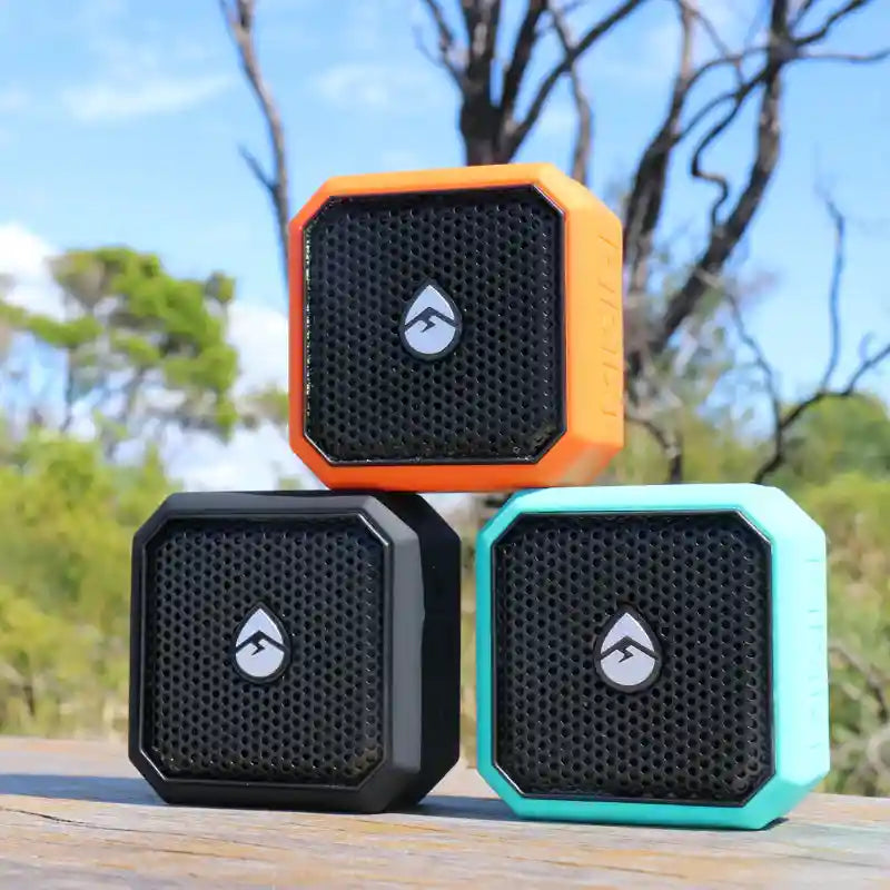 Three portable speakers in orange, black, and teal on a wooden surface with a natural background.