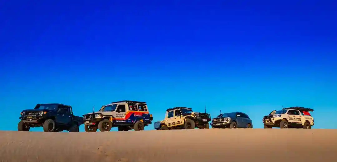 Five off-road vehicles parked on a sand dune with a clear blue sky.