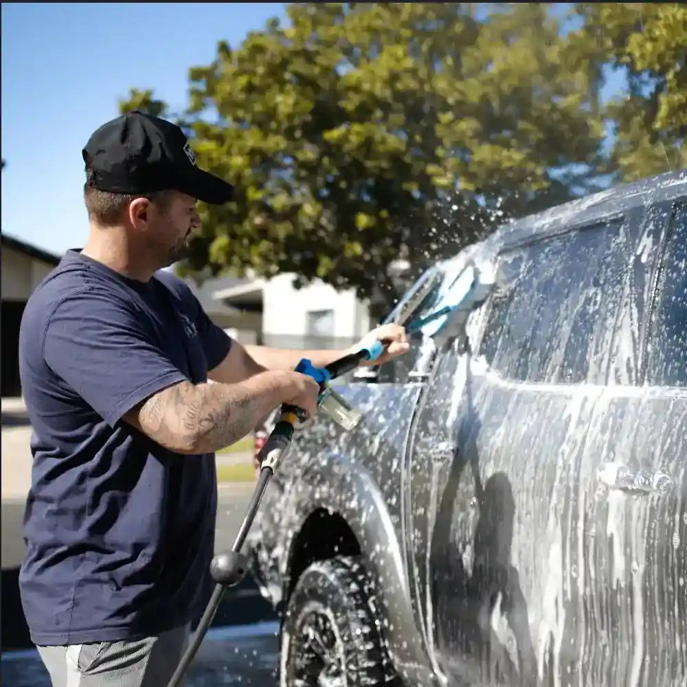 Man washing a car with a sponge and brush, surrounded by trees and clear sky.