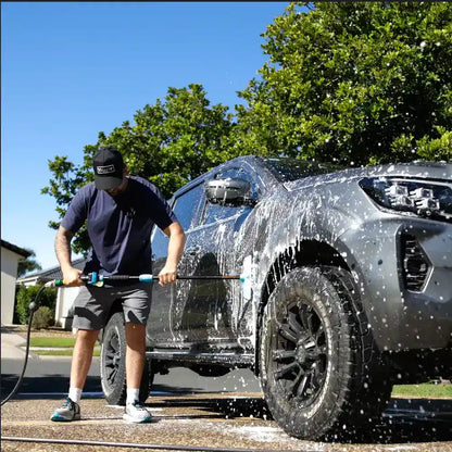 Person washing a gray SUV with a pressure washer on a sunny day.