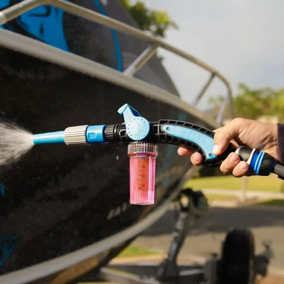 Person using a pressure washer to clean a vehicle with a pink detergent container attached.