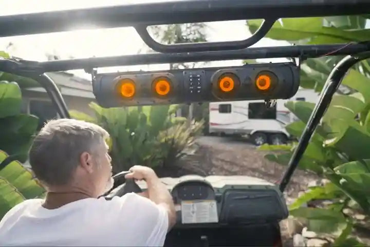 Man driving a vehicle with a camper in the background, surrounded by greenery.