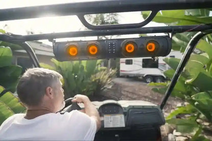 Man driving a vehicle with a camper in the background, surrounded by greenery.